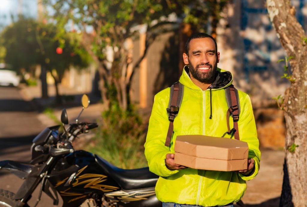 Homem segurando caixa de pizzas com uma moto ao fundo